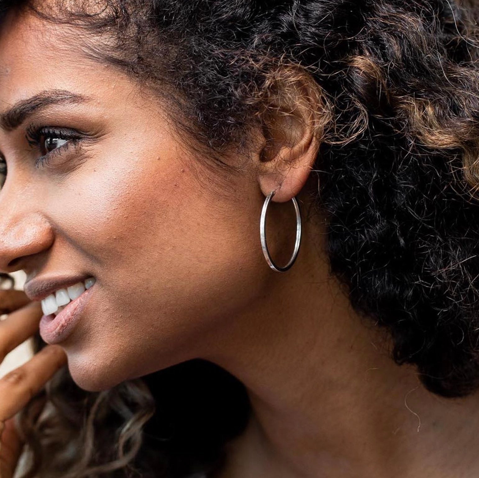 A model with brown curly hair looking left and smiling wearing sterling silver hoop earrings and wearing a white tank top