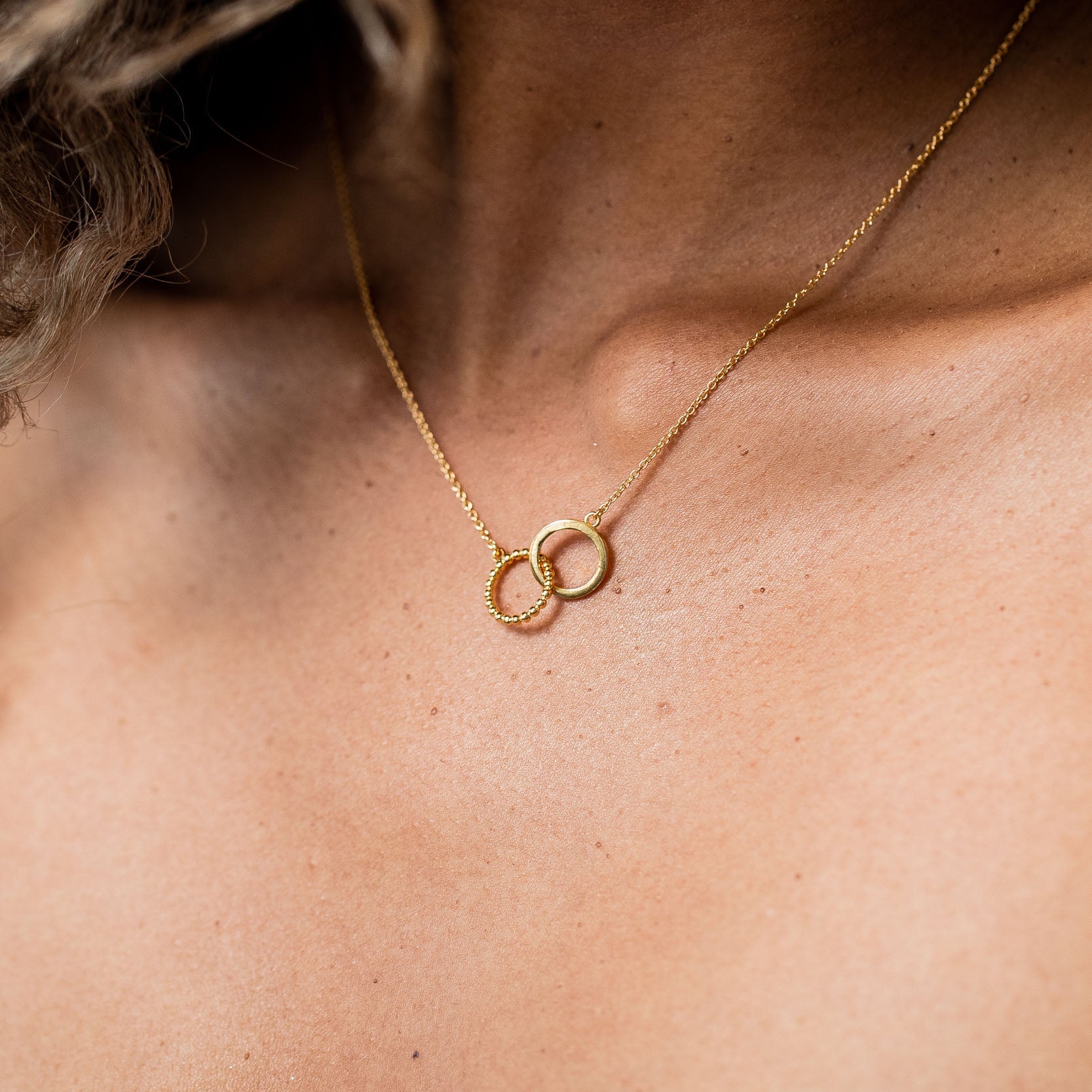 A model with curly blonde and brown hair and light brown skin wearing a gold vermeil necklace with two interlocking circles