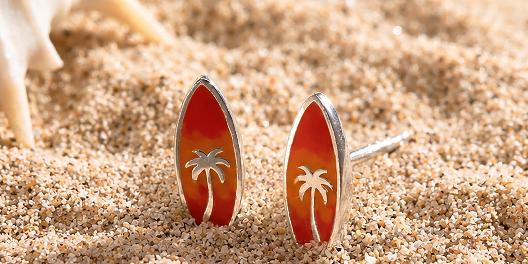 Boma Jewelry Surfboard-shaped earrings with palm tree design on a sandy surface