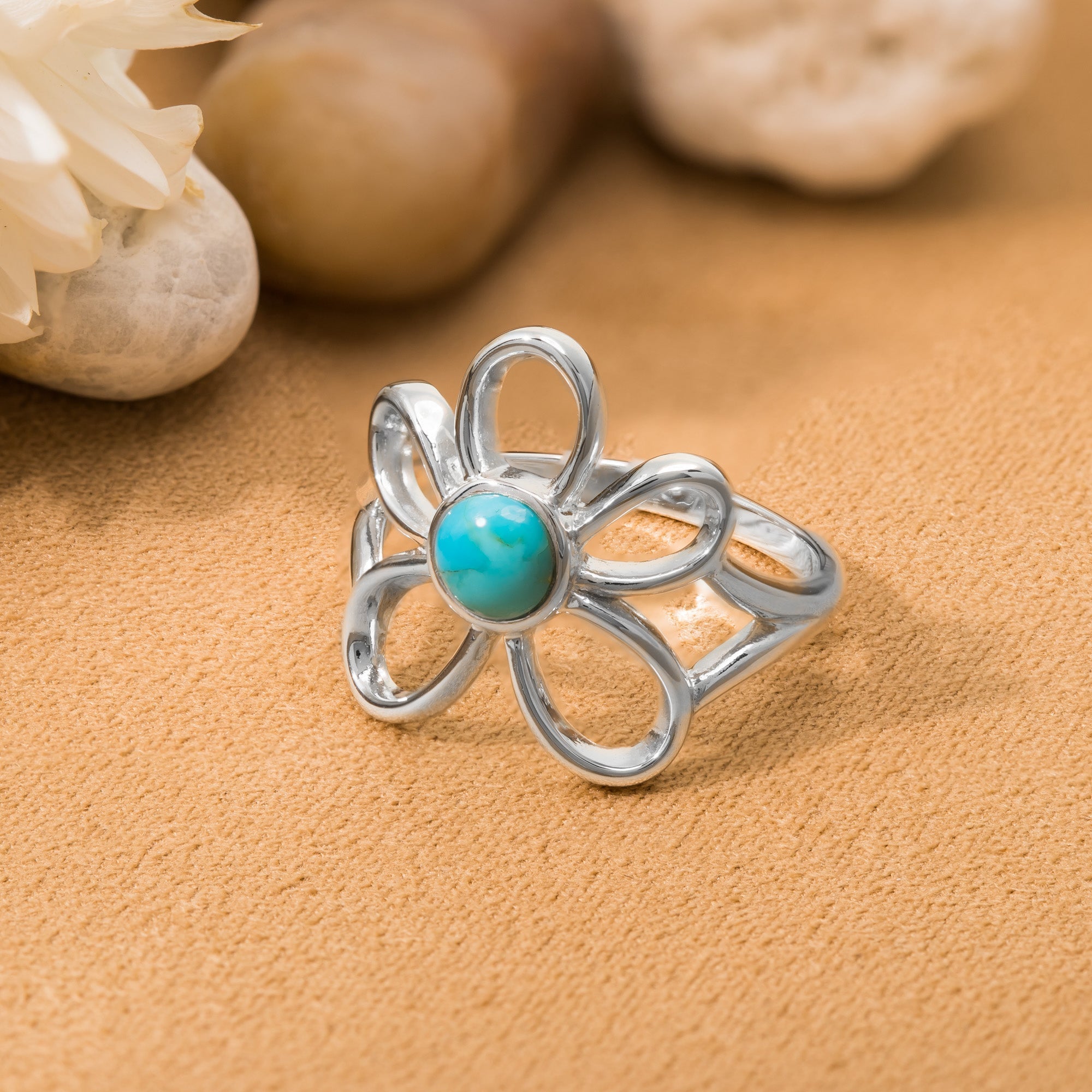 Silver flower-shaped ring with an open petal design and a round turquoise stone at the center, displayed on a warm sandy-colored surface with softly blurred natural elements in the background.