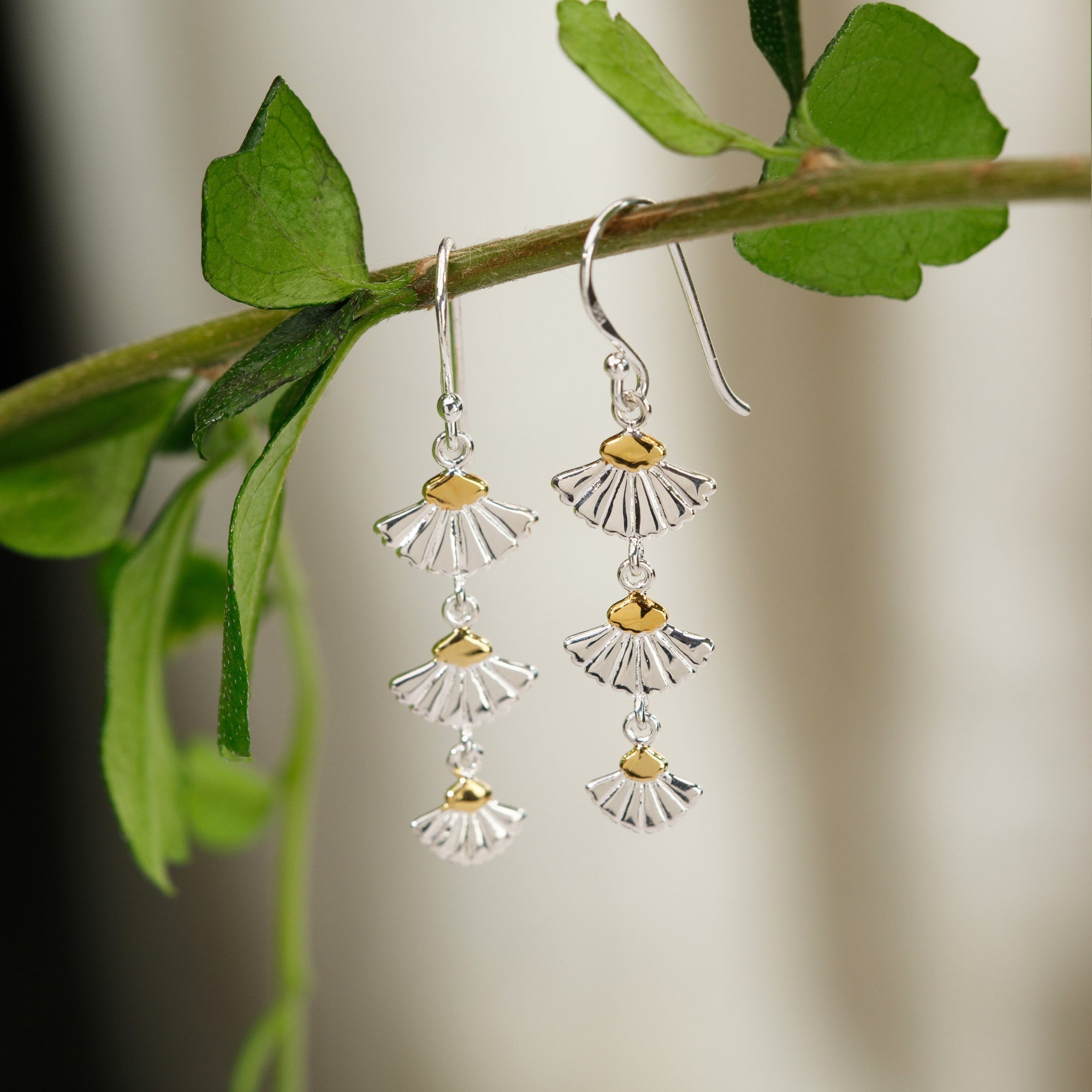 **Alt text:** Silver and gold-tone tiered fan dangle earrings hanging from a green leafy branch, featuring delicate cascading fan-shaped elements against a softly blurred natural background.