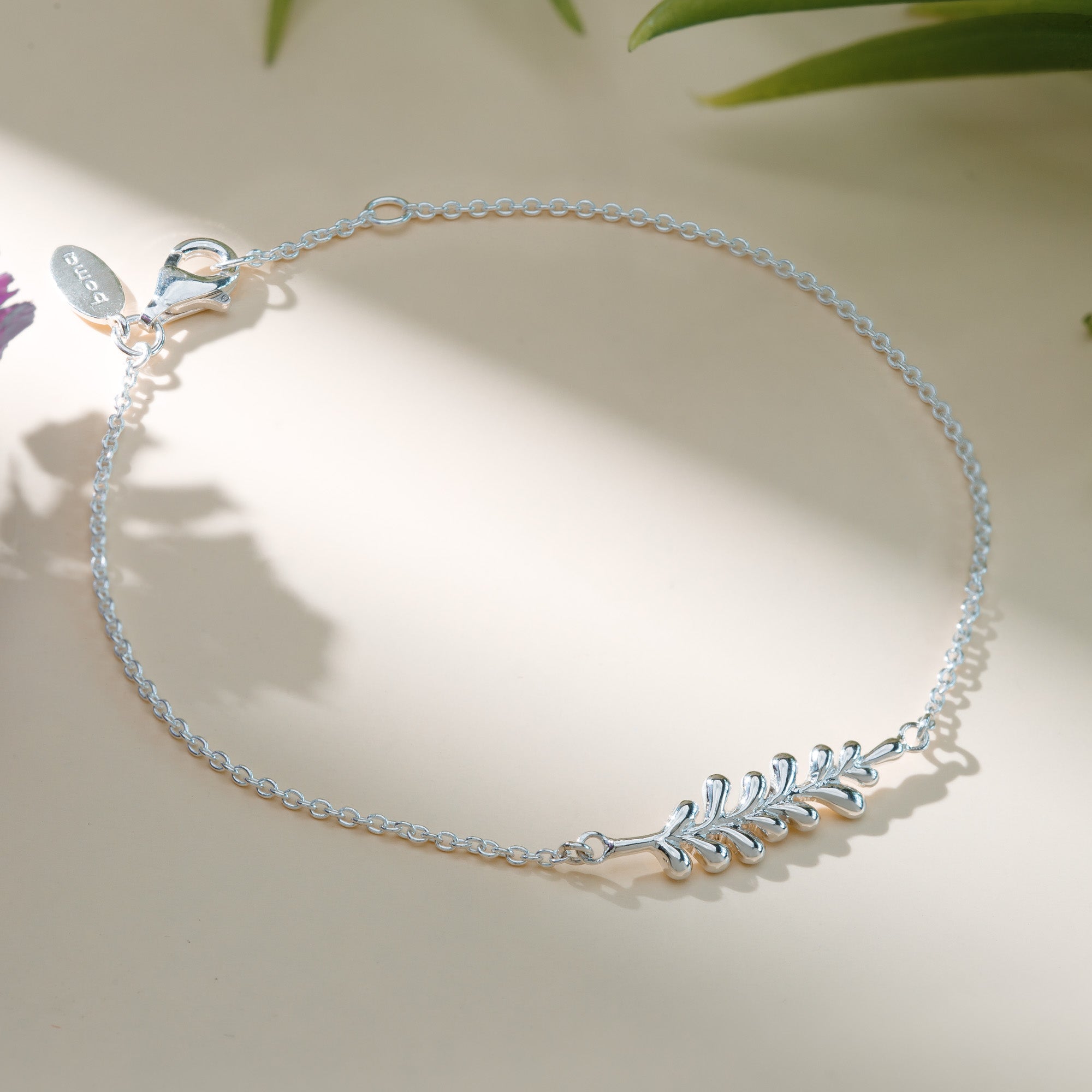 Delicate sterling silver bracelet featuring a small leaf branch motif at the center, displayed on a soft cream background with natural light and gentle botanical shadows.